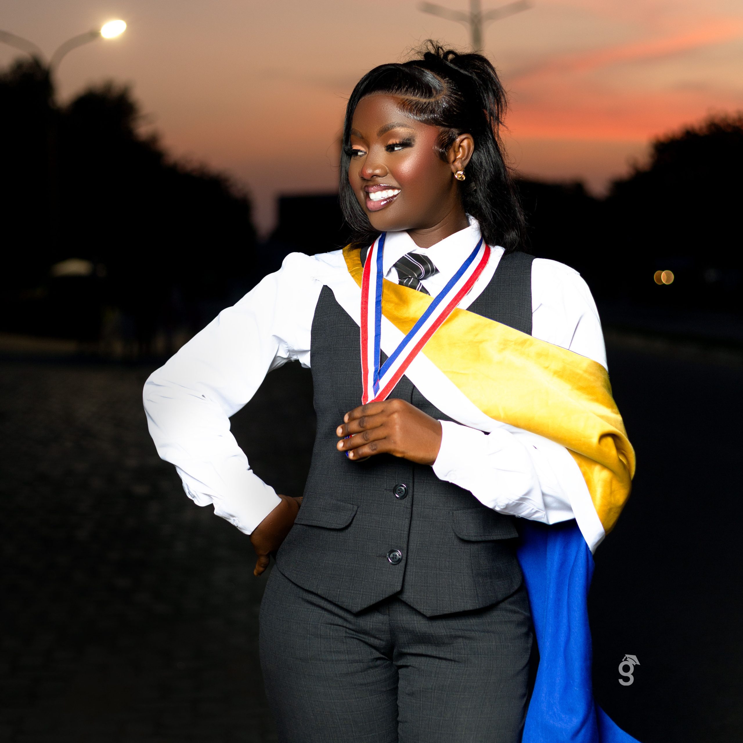 Gradmoment sunset graduation portrait of a student in a waistcoat and yellow sash holding her medal