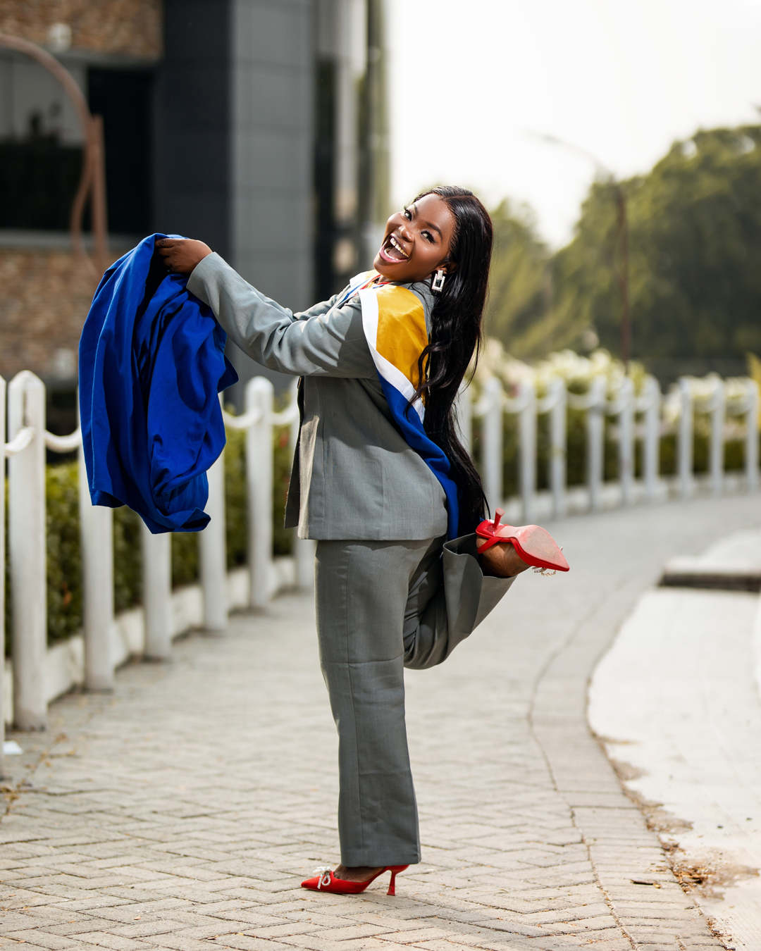 A joyful graduate in a grey suit and yellow sash holding her blue academic gown outdoor at the UEW Student Center
